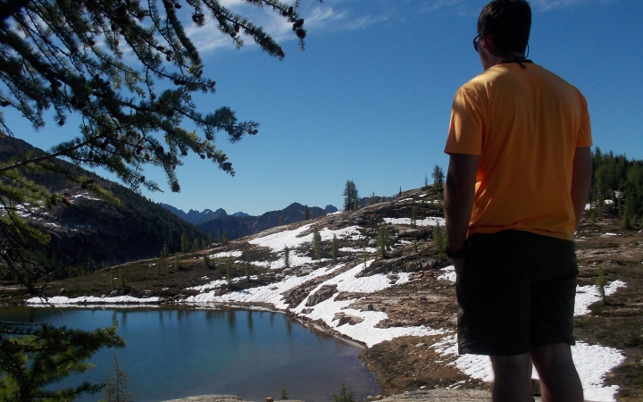 A person faces away from the camera, looking out over a small body of water and a landscape dotted with snow.